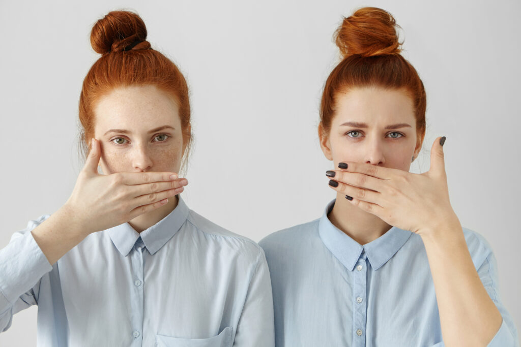 Portrait of two attractive redhead women in identical shirts covering lips with hands, having common secret, forbidden to say anything, posing indoors at white studio wall with serious looks