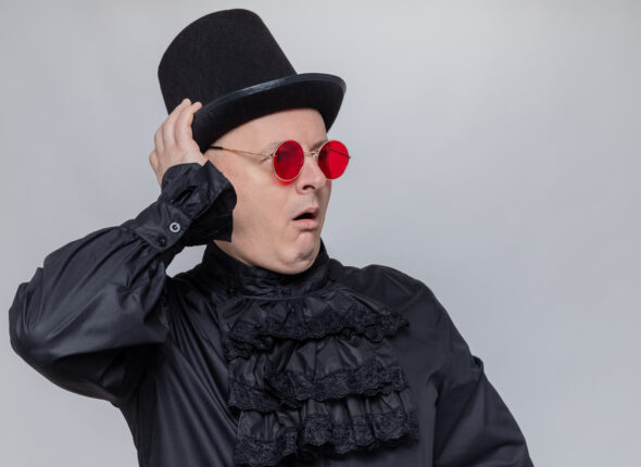 excited adult slavic man with top hat and with sunglasses in black gothic shirt looking at side isolated on white background with copy space