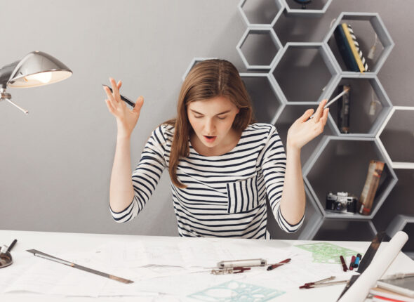 OMG. Portrait of young unhappy beautiful young brow-haired female engineer spreading hands with opened mouth, noticed mistake in drawings she made. Body language.