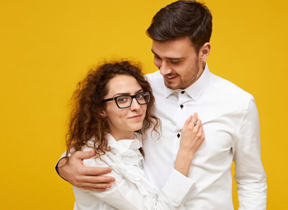 Portrait of timid young female with curly dark hair looking at camera with shy smile embracing her tall boyfriend with stubble. Handsome guy going to kiss his charming girlfriend, being in love