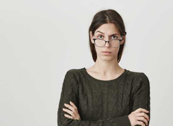 Indoor portrait of young woman in spectacles has serious confident expression, keeps arms crossed, feels angry after quarrel with colleagues. European irritated female looks dissatisfied