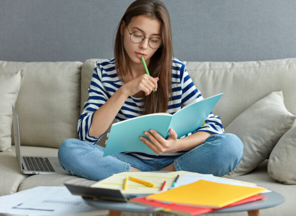 Studying online concept. Serious young woman being busy with remote freelance project, sits at comfortable sofa, writes notes, holds textbook, use laptop computer at home with wireless internet